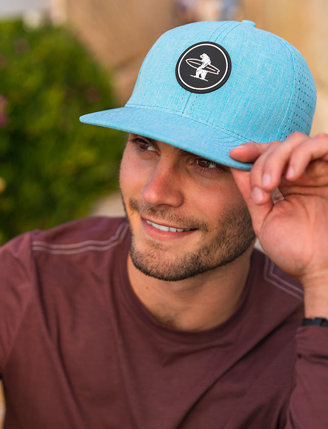 Male model wearing a light blue snapback hat with a logo of a white bear holding a white surfboard on the front.