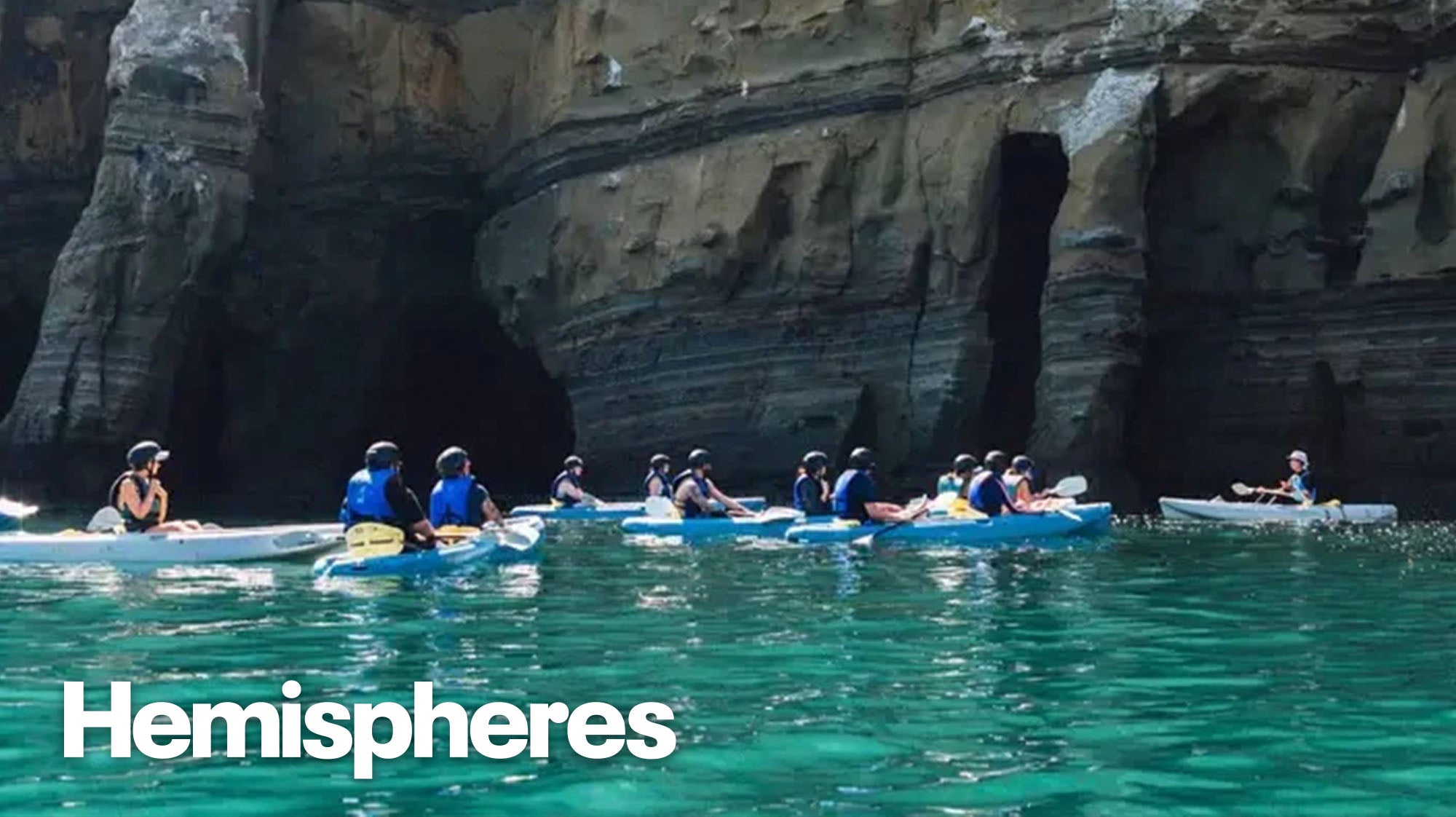 A group of kayakers in the open ocean in front of a series of sea caves.