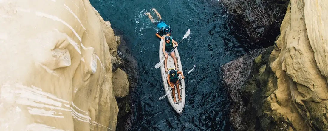 Two kayakers in a single kayak being guided into one of the Seven Sea Caves in La Jolla by an Everyday California kayak guide who is swimming in the water behind them.